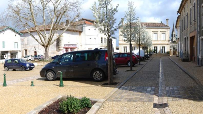 Réaménagement sobre des espaces publics de Gensac. Grâce au traitement de sol, la voiture n'apparait pas prioritaire, tout en ayant sa place en ville. Visuellement le parking est intégré au traitement général de la place, sans être trop différencié, les arbres replantés suffisent à délimiter les stationnements sans marquages au sol, le trottoir à droite n'est pas encombré de bornes anti-stationnement